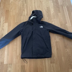 Svart vindjacka från The North Face - Säljer en svart vindjacka från The North Face i storlek S. Jackan har huva, dragsko nertill och klassisk logga på bröstet och ryggen. 