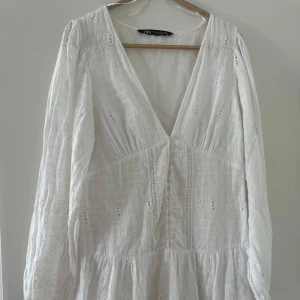 Vit broderad klänning från Zara - Superfin vit miniklänning från Zara med broderade blommor. Klänningen har v-ringning, knappar framtill och långa ärmar. 