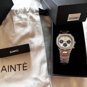 Silverfärgad klocka från Cainte - Stilren klocka från Cainte med silverfärgat armband i metall och vit urtavla med tre svarta subdials. Klockan har en modern, åttakantig boett och detaljerad urtavla med mönster. Perfekt accessoar för dig som gillar en clean och exklusiv look.