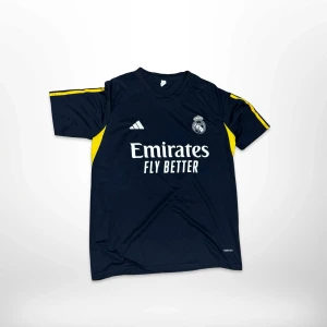 Real Madrid marinblå fotbollströja Adidas - Snygg marinblå Real Madrid fotbollströja från Adidas med vit logga och tryck på bröstet. Tröjan har gula paneler på sidorna och gula detaljer på ärmarna, samt lila och vita ränder på ena ärmen. Tillverkad i lätt och ventilerande polyester.