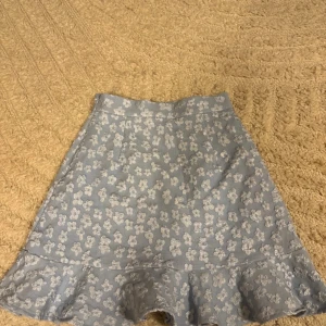 Blommig blå volangkjol från Pieces - Supersöt ljusblå kjol från Pieces med vita blommor och volangkant längst ner. Kjolen har en feminin A-linjeform och är perfekt för dig som gillar romantiska detaljer. Materialet känns mjukt och har en lätt struktur som ger extra stilpoäng.