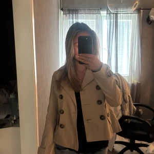 Beige kort trenchcoat med knappar - Säljer en snygg beige trenchcoat med dubbelknäppning och stora bruna knappar. Jackan har klassisk krage, långa ärmar med knappdetalj vid mudden och två fickor framtill. Perfekt för dig som vill ha en stilren och tidlös look i vår eller höst.