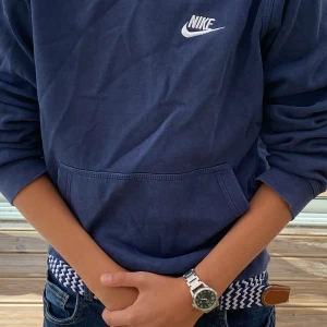 Mörkblå Nike hoodie med huva - Säljer en mörkblå hoodie från Nike med klassisk logga broderad i vitt på bröstet. Tröjan har huva, känguruficka och långa ärmar. Perfekt för dig som gillar en sportig och avslappnad stil. Materialet känns mjukt och skönt mot huden.