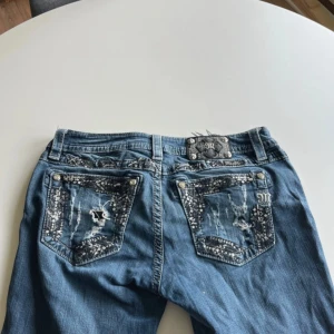 Miss Me jeans med broderade detaljer - Snygga blå jeans från Miss Me med unika broderade och slitna detaljer på bakfickorna. Jeansen har silverfärgade nitar och dekorativa sömmar, samt en cool patch med logga bak i midjan. Klassisk femficksmodell i bomullsmix med riktigt statement-vibe.