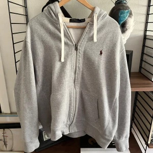 Grå hoodie från Polo Ralph Lauren XL - Klassisk grå hoodie från Polo Ralph Lauren i storlek XL. Tröjan har dragkedja, huva med vita snören och den ikoniska broderade loggan på bröstet. Tillverkad i mjuk bomullsmix med känguruficka framtill. Perfekt för en chill och stilren look.