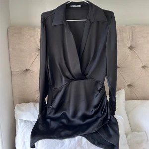 Svart omlottklänning i satin - Snygg svart kort omlottklänning från Zara i glansigt satinmaterial. Klänningen har lång ärm, djup v-ringning och krage som ger en cool och dressad vibe. Perfekt för dig som gillar stilrena och trendiga plagg med lite edge.