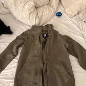 Olivgrön hoodie från Canada Goose - Säljer en olivgrön hoodie från Canada Goose med dragkedja och huva. Tröjan har två stora fickor framtill och en broderad patch på ärmen. Perfekt för dig som gillar streetwear och vill ha något bekvämt och snyggt.