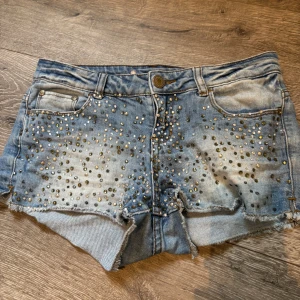 Blå jeansshorts med paljetter - Snygga blå jeansshorts från Met Jeans med råa kanter och massor av glittriga paljetter på framsidan. Shortsen har fem fickor, slitningar bak och en cool tvättad look. Perfekta för dig som gillar att sticka ut och vill ha något extra till sommaren.
