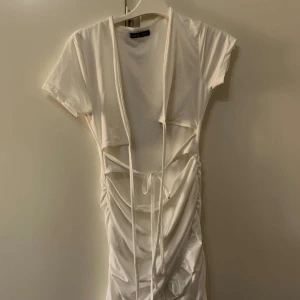 Vit omlottklänning från Shein - Säljer en vit kortklänning från Shein med coola snörningsdetaljer och omlott-design i midjan. Klänningen har korta ärmar och snygga draperingar som ger en trendig look. Perfekt för dig som gillar att sticka ut med din stil.