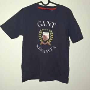 Mörkblå GANT t-shirt med tryck - Snygg mörkblå t-shirt från GANT med klassiskt tryck framtill med sköld, stjärnor och lagerkrans. Rund halsringning med röd detalj på insidan. Perfekt för dig som gillar preppy stil och vill ha något bekvämt och stilrent.
