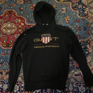 Svart hoodie från Gant XS - Svart hoodie från Gant i storlek XS med broderad logga och texten 'American Sportswear' framtill. Tröjan har huva med snörning och klassisk collegekänsla. Perfekt för dig som gillar sportig och stilren look.
