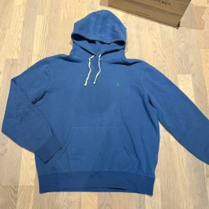Blå Ralph Lauren hoodie - Säljer en blå hoodie från Polo Ralph Lauren med klassisk grön broderad logga på bröstet. Storlek L. Han på bild är 185cm och väger 74kg. Perfekt för dig som gillar en avslappnad och stilren look.