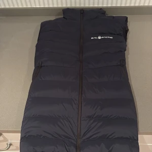 Svart dunväst från Sail Racing - Säljer en svart dunväst från Sail Racing med hög krage och dragkedja framtill. Västen har två sidofickor med dragkedja och logga på bröstet samt ryggen. Perfekt för lager-på-lager och håller dig varm under kyliga dagar.