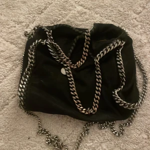 Svart axelväska Stella McCartney - Säljer en svart axelväska från Stella McCartney med grova silverfärgade kedjor och rund logotypbricka. Väskan är tillverkad i syntetmaterial och har en cool, modern look. Perfekt för dig som vill ha en statementväska med lyxig känsla. Kan även tänka mig att gå ner pris vid snabb affär! 