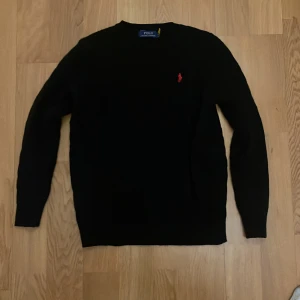 Svart stickad tröja Polo Ralph Lauren - Klassisk svart stickad tröja från Polo Ralph Lauren med röd broderad logga på bröstet. Tröjan har rund halsringning och långa ärmar, perfekt för dig som gillar stilren och tidlös design.