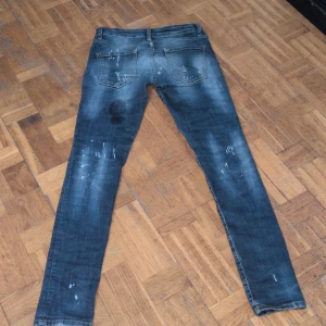 Blå slitna skinny jeans med hål - Säljer ett par blå skinny jeans med coola slitningar, hål och färgstänk för en riktigt edgy look. Jeansen har låg midja och klassisk femficksdesign. Perfekta för dig som gillar streetstyle och vill sticka ut lite extra.