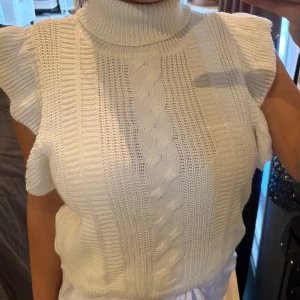 Vit stickad ärmlös polotröja - Supersnygg vit stickad polotröja med ärmlös design och volangdetaljer vid axlarna. Tröjan har ett klassiskt flätmönster framtill och hög krage som ger en trendig look. Perfekt att styla med jeans eller kjol för en chic vibe.