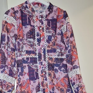 Mönstrad långärmad klänning med spets - Färgglad långärmad klänning med patchworkmönster i lila, rosa och orange toner. Klänningen har vita spetsdetaljer och knappar framtill samt dekorativa band längs ärmar och midja. Lätt och luftig känsla, perfekt för dig som gillar boho-vibe. Obs! Storleken kan passa XS/S och M