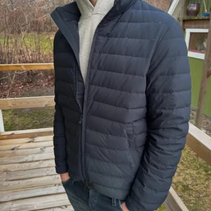 Svart pufferjacka med dragkedja - Snygg svart pufferjacka med horisontella sömmar och hög krage. Jackan har en stilren look och stängs med dragkedja framtill. Perfekt för kyliga dagar och passar till det mesta i garderoben.