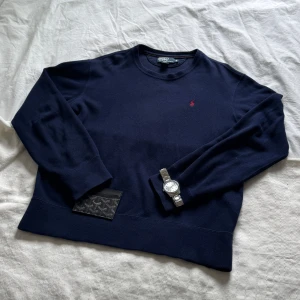 Mörkblå tröja från Polo Ralph Lauren - Klassisk mörkblå tröja från Polo Ralph Lauren med rund halsringning och diskret röd logga broderad på bröstet. Tröjan har långa ärmar och är tillverkad i mjuk bomull, perfekt för en stilren och avslappnad look.