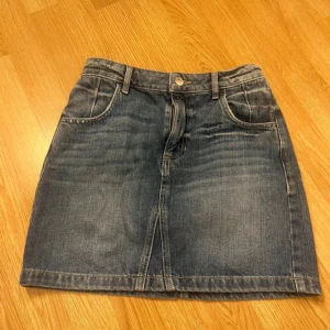 Jeanskjol från Guess med fickor - Snygg klassisk jeanskjol från Guess i mörkblå denim med fyra fickor och bälteshällor. Kjolen har en kort, rak passform och ikonisk Guess-logga på ena bakfickan. Perfekt för dig som gillar en enkel och cool stil. Storlek 26