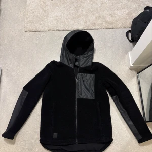 Svart teddyjacka med huva och detaljer från Didrikson - Svart teddyjacka med snygga svarta detaljer i syntet på huva, bröstficka och ärmar. Jackan har dragkedja framtill och en modern, lite längre passform baktill. Perfekt för dig som gillar stilren och sportig look.