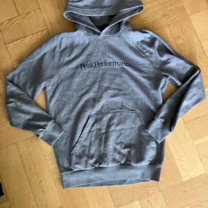 Grå hoodie från Peak Performance - Snygg grå hoodie från Peak Performance med svart logga på bröstet. Klassisk känguruficka framtill, huva och mjukt fleecematerial på insidan. Perfekt för chill dagar eller när du vill ha en enkel och stilren look.
