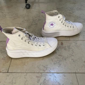 Converse All Star vita sneakers med lila detaljer - Snygga Converse All Star sneakers i vitt canvas med lila detaljer på baksidan och logga. Skorna har hög platåsula, snörning och rund tå. Perfekta för dig som vill sticka ut med klassisk stil och cool twist.