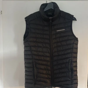 Svart dunväst från Peak Performance - Svart dunväst från Peak Performance med quiltad design och hög krage. Västen har dragkedja framtill och två sidofickor. Perfekt för lager-på-lager och har en clean look med diskret logga på bröstet. Passar dig som gillar sportig och stilren vibe.
