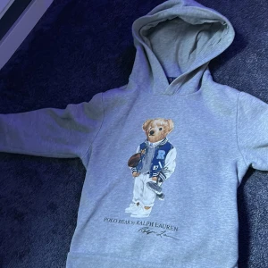 Grå hoodie Polo Bear Ralph Lauren - Snygg grå hoodie från Ralph Lauren med ikoniska Polo Bear-trycket framtill.Storlek 10-12 år. Tröjan har huva som är gjord i mjuk bomullsmix. En liten defekt på trycket men de är inget man märker på avstånd.