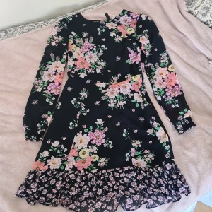 Blommig svart kortklänning med volang - Supersöt svart klänning med färgglatt blommönster i rosa, lila, vitt och grönt. Klänningen har långärmade ärmar med smock vid handlederna, volangdetaljer längs nederkanten och en öppen rygg med volangkant. Perfekt för dig som gillar romantisk och lekfull stil.