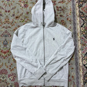 Ljusgrå hoodie från Polo Ralph Lauren - Snygg ljusgrå hoodie från Polo Ralph Lauren med dragkedja och klassisk logga broderad på bröstet. Tröjan har huva med snörning, långa ärmar och två fickor framtill. Perfekt för en chill och stilren look.