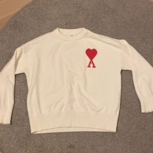 Vit stickad tröja från Ami Paris - Vit stickad tröja från Ami Paris med röd hjärtlogga och bokstaven A på bröstet. Tröjan har rund hals, ribbade muddar och är långärmad. Perfekt för dig som gillar stilrena plagg med en twist.