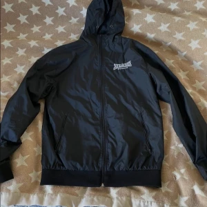 Svart vindjacka från Sofiläkaren - Svart vindjacka med huva från Sofiläkaren Stockholm. Jackan har dragkedja framtill, två fickor med dragkedja och ribbade muddar vid ärmslut och nederkant. Perfekt för blåsiga dagar och enkel att matcha med det mesta.