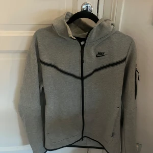 Grå Nike hoodie med dragkedja - Snygg grå hoodie från Nike med svart logga på bröstet och hel dragkedja framtill. Tröjan har huva, långa ärmar och svarta detaljer längs dragkedjan och sömmarna. Perfekt för chill eller träning, med fickor på sidorna och en extra ficka på ärmen.