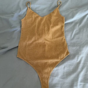 Glittrig guld top från H&M - Top från H&M i glittrigt guld med smala axelband och v-ringad front. Toppen har en high-cut trosa och en topp med öppen rygg. Materialet är mjukt 