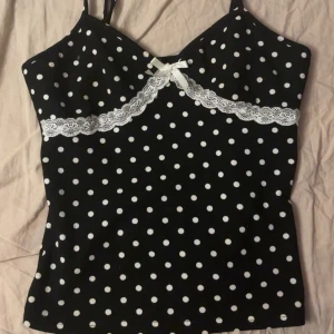 Svart prickig linnetopp med spets - Supersöt svart linnetopp med vita prickar, spetsdetalj och liten rosett framtill. Smala axelband och v-ringad skärning. Perfekt för en söt  look.