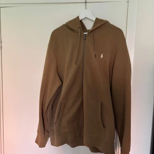 Beige hoodie från Polo Ralph Lauren - Säljer en beige hoodie från Polo Ralph Lauren med klassisk broderad logga på bröstet. Tröjan har huva med snörning, dragkedja framtill och två stora fickor. 