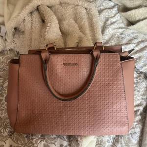 Rosa handväska från Vezzano - Säljer en snygg rosa handväska från Vezzano med präglat mönster och guldfärgade detaljer. Väskan har både handtag och axelrem samt dragkedja upptill och ett extra fack med dragkedja på baksidan. Perfekt storlek för allt du behöver ha med dig.