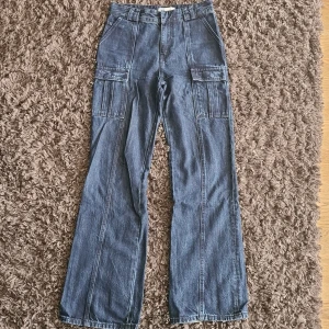 Blå cargopants i jeans från Zara - Snygga blå cargopants från Zara i jeansmaterial. Byxorna har hög midja, breda ben och flera stora fickor på sidorna samt bakfickor med dragkedja. Perfekta för en cool och avslappnad stil. Passar dig som gillar streetwear och praktiska detaljer.