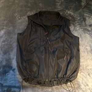 Svart väst från The Best med snörning - Svart väst från The Best med dragkedja framtill, flera fickor och justerbar snörning i nederkant. Västen har en enkel, sportig look och hög krage. Perfekt för lager-på-lager och chill stil.