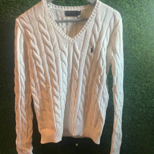 Vit kabelstickad tröja Polo Ralph Lauren - Vit kabelstickad tröja från Polo Ralph Lauren med klassisk v-ringning och broderad logga på bröstet. Tröjan har långa ärmar och ribbade muddar vid hals, ärmslut och nederkant. Perfekt för dig som gillar preppy stil och vill ha något som känns både chill och snyggt.