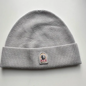 Grå mössa från Parajumpers - Snygg grå mössa från Parajumpers med uppvikt kant och broderad logga framtill. Tillverkad i mjuk och varm stickad akrylblandning, perfekt för kyliga dagar. Enkel design som passar till de flesta stilar och jackor.
