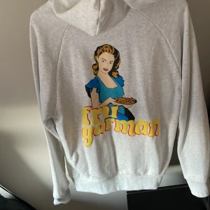Ljusgrå hoodie Fru Garman Revenite M - Ljusgrå hoodie från Revenite i storlek M med tryck på ryggen av en kvinna och texten 'Fru Garman' i färgglada bokstäver. Tröjan har huva med snörning, magficka och långa ärmar.