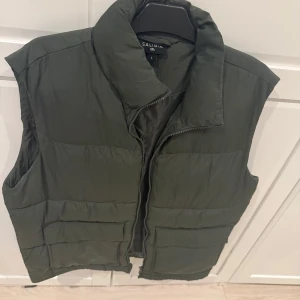 Mörkgrön dunväst från Saligia - Snygg mörkgrön dunväst från Saligia med hög krage och dragkedja framtill. Västen har en quiltad look och är perfekt att slänga över en hoodie eller tröja. Enkel och stilren design som funkar till många outfits.