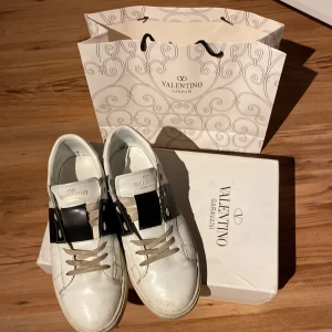 Valentino Garavani Open sneakers vit/svart - Snygga Valentino Garavani Open sneakers i vitt skinn med bred svart läderdetalj över vristen och beige snörning. Klassisk rund tå och platt sula. Kommer med originalkartong och påse. Perfekta för dig som vill ha en lyxig och stilren look.