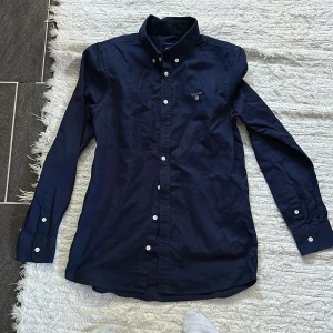 Mörkblå skjorta från GANT - Klassisk mörkblå linne skjorta från GANT med vit broderad logga på bröstet. Skjortan har lång ärm, knappar framtill och på manschetterna samt button down-krage. Tillverkad i mjuk bomull, perfekt för en clean och stilren look.