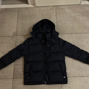 Blå pufferjacka från Polo Ralph Lauren - Säljer en svart pufferjacka från Polo Ralph Lauren med huva och dragkedja. Jackan har en klassisk pufferlook med breda kanaler och den ikoniska röda loggan på bröstet. Perfekt för kalla dagar och har praktiska fickor med dragkedja.