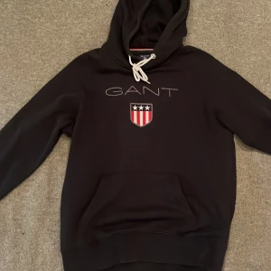 Svart hoodie från GANT med logga - Svart hoodie från GANT med stor broderad logga och sköld på bröstet. Tröjan har huva med vita snören och en stor magficka. Perfekt för dig som gillar klassisk streetstyle och vill ha något bekvämt och snyggt.