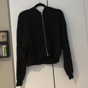 Svart hoodie med dragkedja från H&M - Svart hoodie från H&M i storlek M med huva och sned dragkedja framtill. Tröjan har långa ärmar, känguruficka och ribbade muddar. Perfekt för en chill och avslappnad stil.
